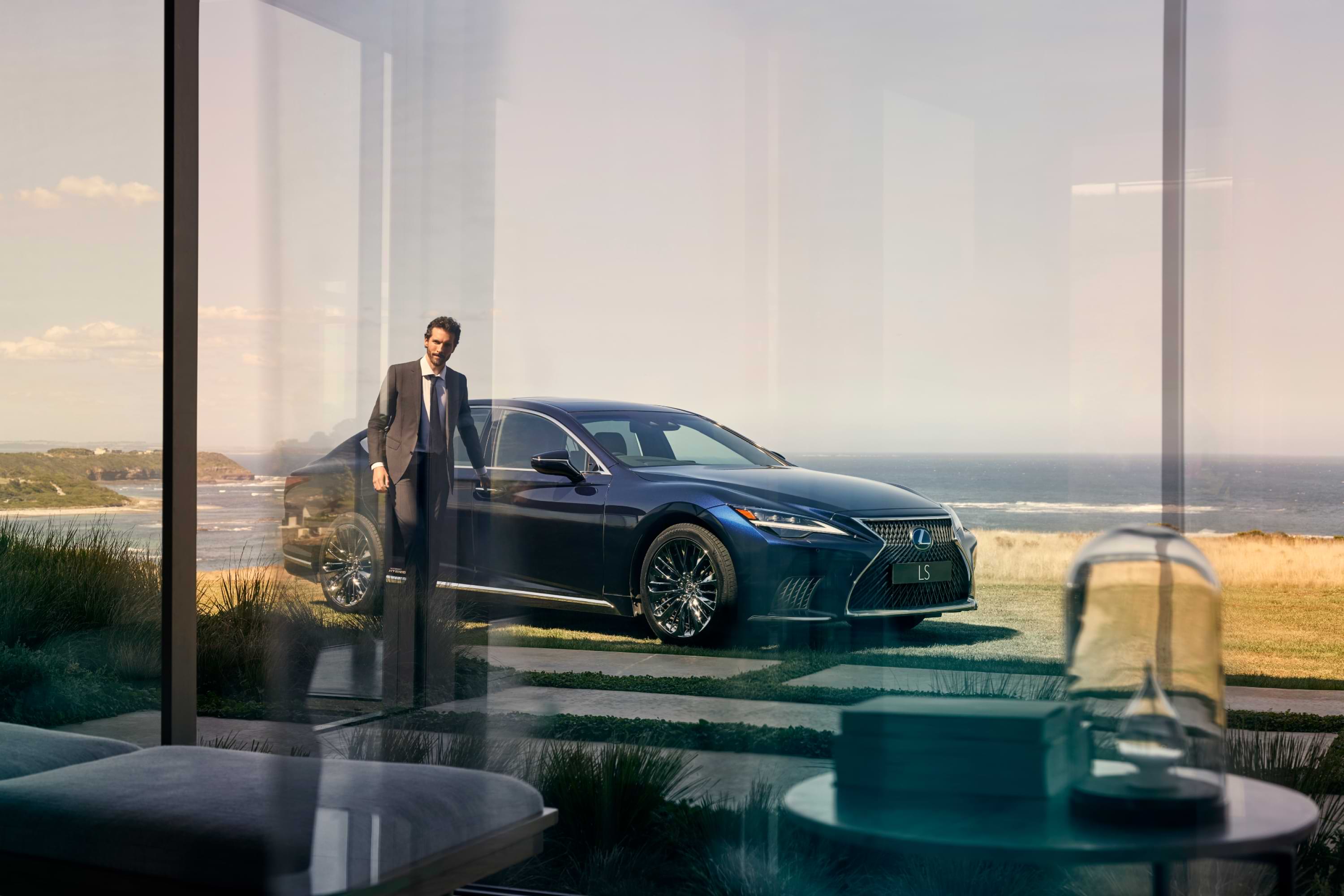 Lexus LS Hybrid hero - man stands with hand on driver&#39;s door handle. He is looking through and into a glass window of a house. The sea is behind them.