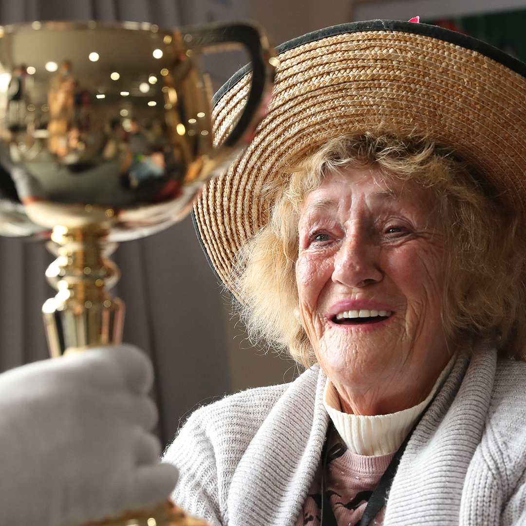 An elderly lady smiles as someone shows her the 2021 Cup. She is wearing a wicker hat.