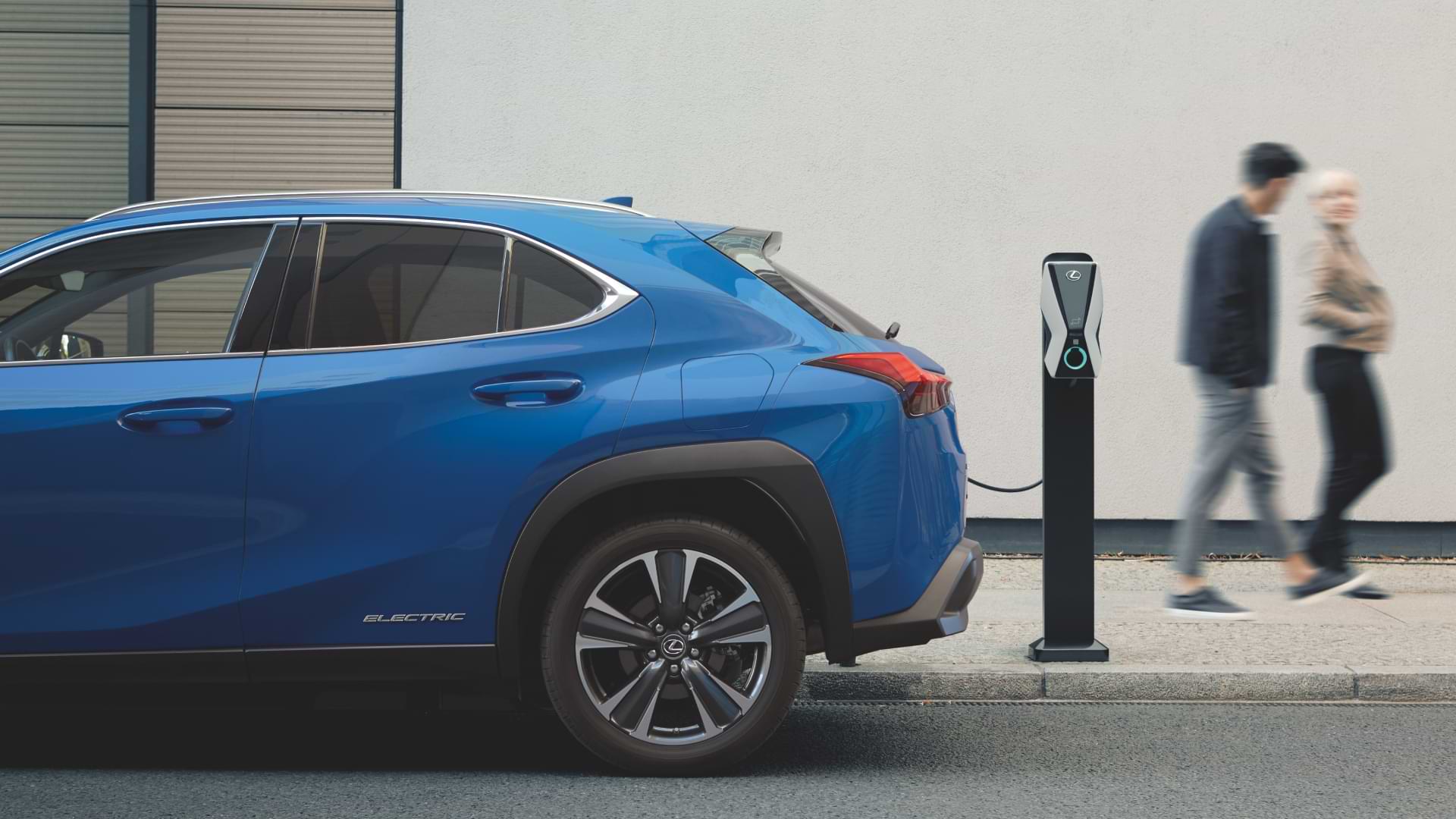 A parked, charging UX 300e sits on the street, two people are seen walking past behind the car, on the footpath.